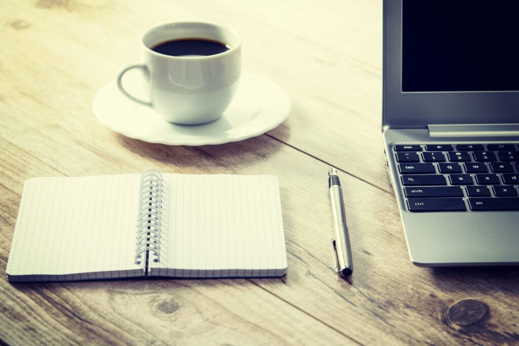 A cup of black coffee, an open notebook, pen and macbook on a wooden desk. In a sepia tone.