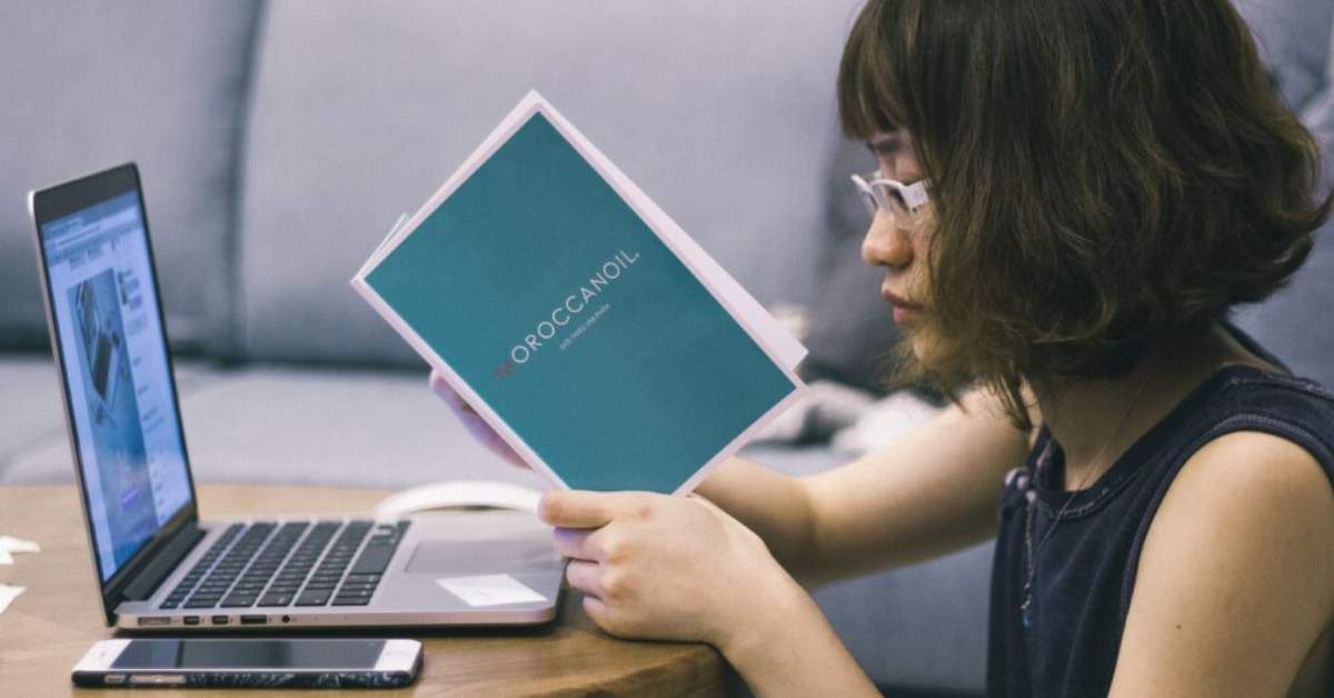 Female sits at a laptop whilst reading a printed booklet