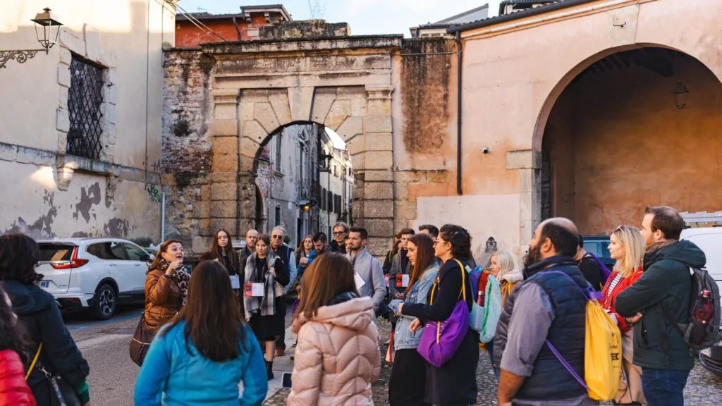 An image of a group of people at an outside activity, as part of an event. The group is stood by the old walls of an Italian city as someone speaks to them.