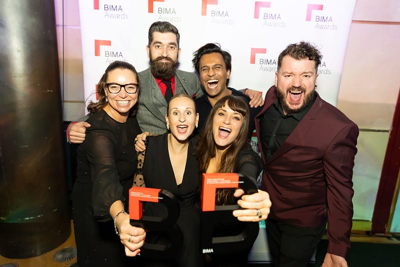 A group of 6 people celebrating their BIMA awards - stood in front of BIMA-themed backdrop, holding their 2 award trophies and smiling