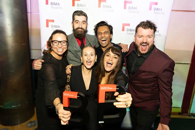 A group of 6 people celebrating their BIMA awards - stood in front of BIMA-themed backdrop, holding their 2 award trophies and smiling