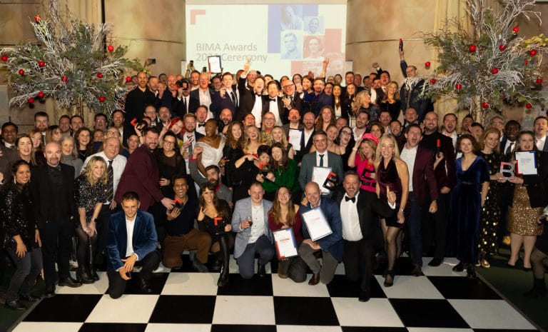 Large group of people at the BIMA Awards ceremony, celebrating with winners holding their award trophies aloft