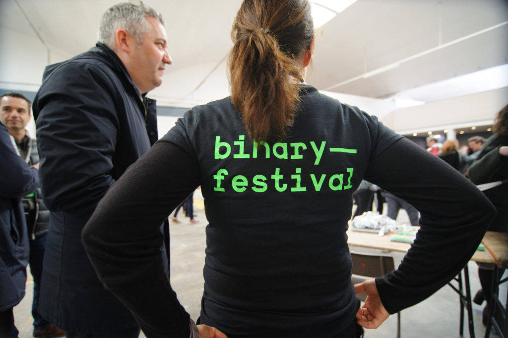 Woman with brown hair in a ponytail, standing with her hands on her hips, with her back to the camera wearing a black t-shirt saying "Binary Festival" on the back in bright green. Next to her on the left is a man with a black coat on. They are both standing at an event venue.