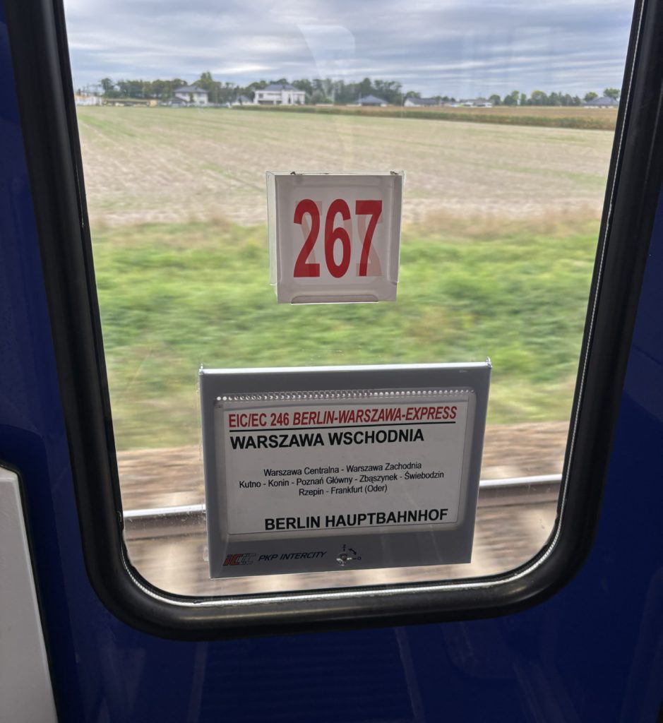 image of farmland framed by a train window, the number 267 on a sticker and the sign says Warsaw Centralna and Berlin Hauptbarnhof