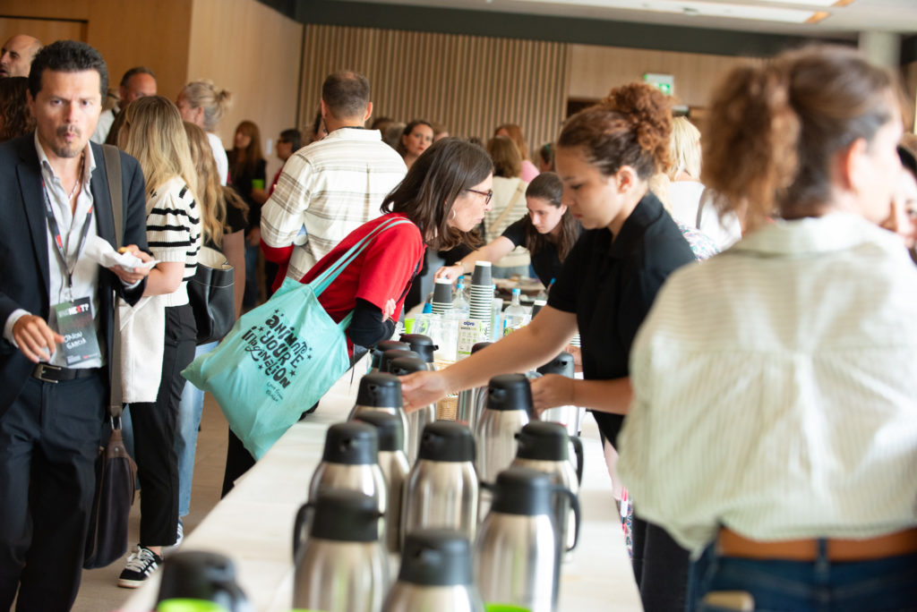 An image of a busy room of people at an event. A central table runs down the middle of the room, covered in tea and coffee jugs. 2 women lean in from either side of the table to make drinks whilst others are chatting in multiple groups around the sides of the room.