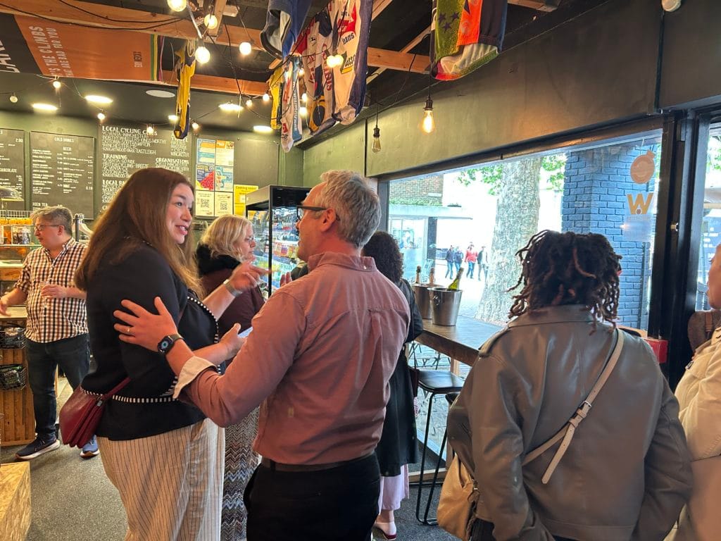 people saying hello at an event in a coffee shop. It shows a woman welcoming a man to the event and a queue behind him with another woman. In the background is the service team getting drinks ready